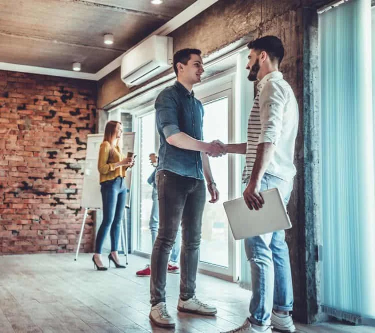 Deux hommes en tenue décontractée se serrent la main dans un bureau moderne, tandis qu'une femme écrit sur un tableau de papier et qu'une autre se tient près d'une fenêtre à l'arrière-plan.