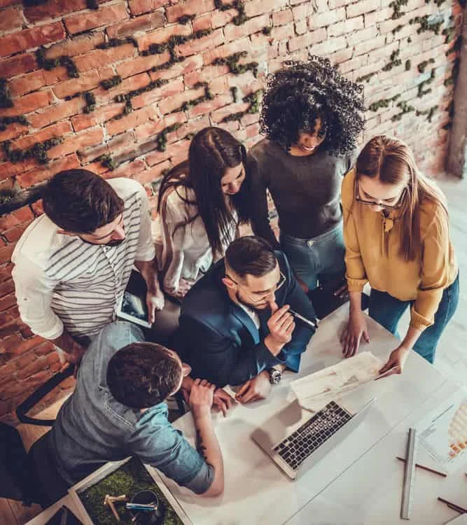 Six personnes réunies autour d'une table, regardant des documents et un ordinateur portable, collaborant dans un bureau moderne avec un mur de briques en arrière-plan.